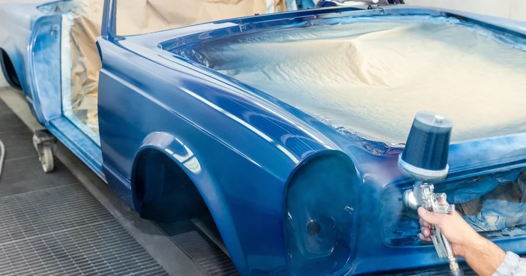 Carrosserie bleue d'une voiture ancienne en cabine de peinture, un pistolet à peinture pulvérise la teinte sur l'aile avant